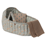 A quilted baby bassinet or carrier seat in a muted sage green floral print, shown with a patterned insert cushion and a folded brown knit blanket on a plain white background.